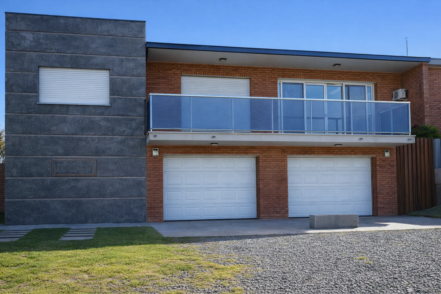 Frente de vivienda de dos plantas con doble garaje, ladrillo visto y balcón corrido con baranda de vidrio.