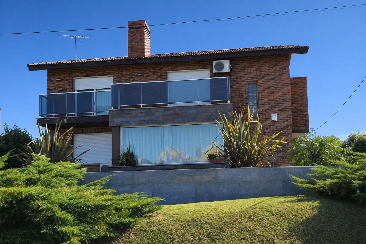 Vista desde el jardín de una casa de ladrillo visto con gran ventanal, balcón superior y parquización al frente.