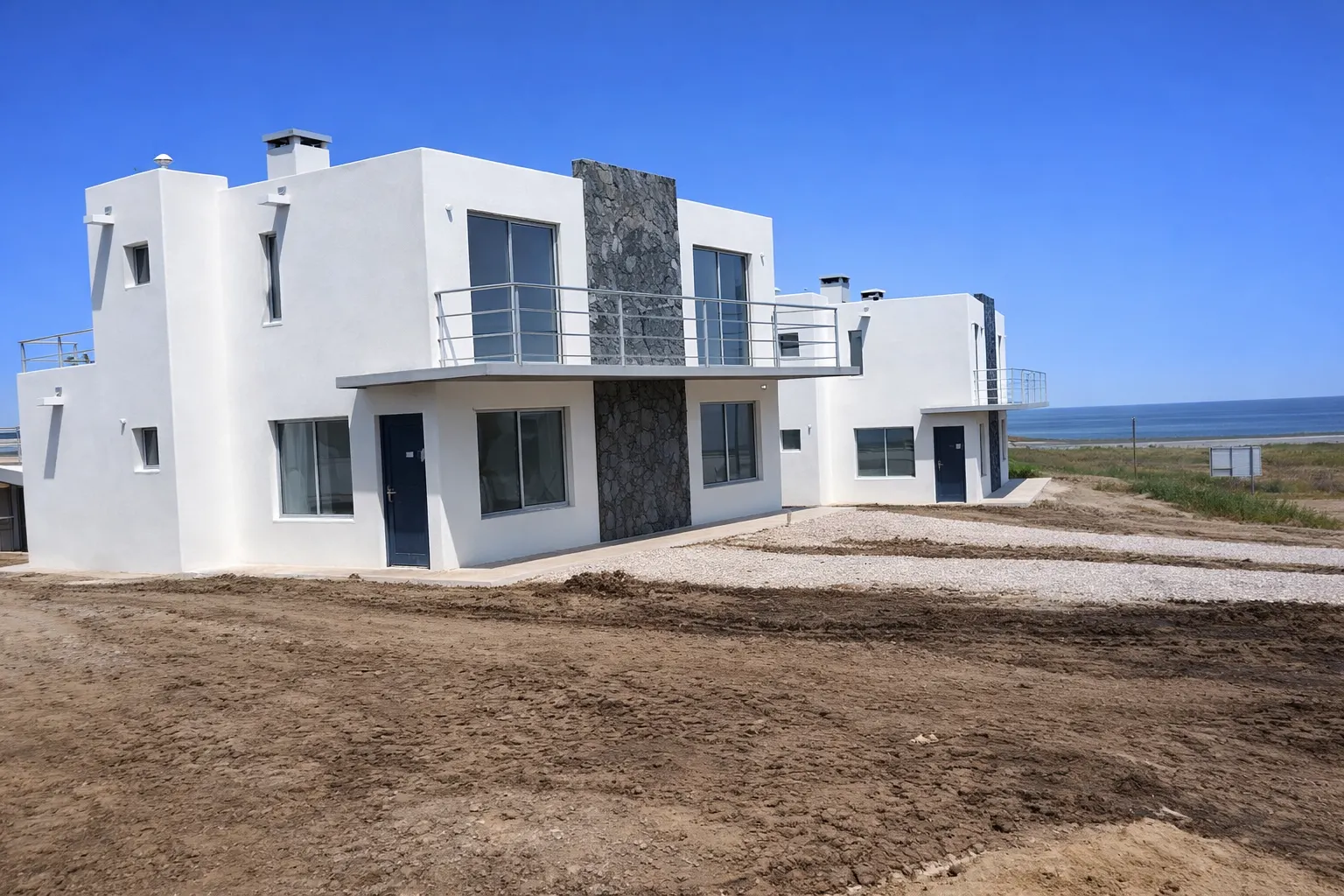 Vista lateral del conjunto con dos bloques terminados y el mar al fondo, mostrando implantación y acceso vehicular.