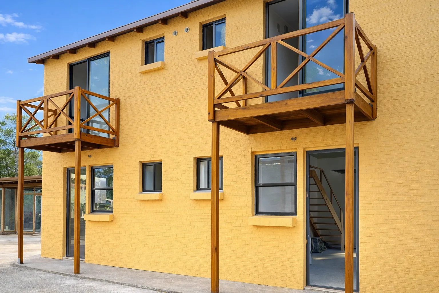 Fachada amarilla de dos plantas con balcones de madera, carpinterías negras y acceso lateral techado.