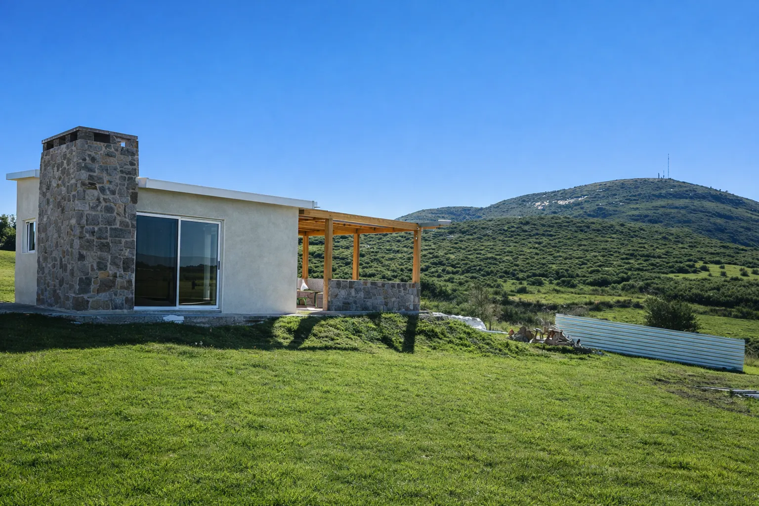Vista lateral de la vivienda con fachada clara, gran paño vidriado y sierras verdes de fondo.